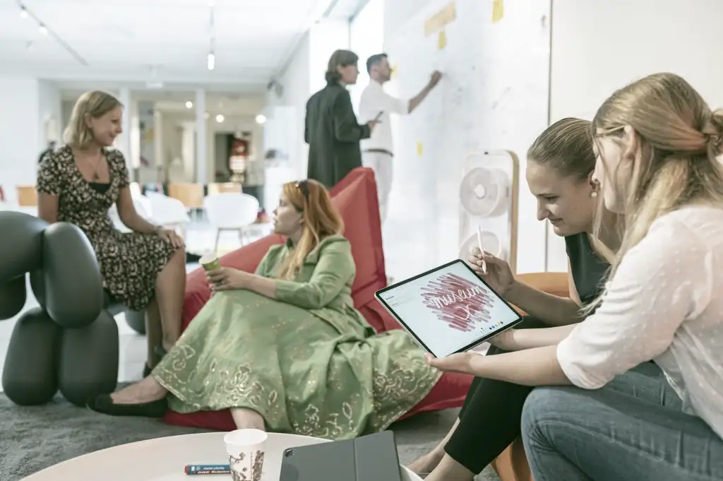 Ein großer, heller Raum. Zwei Frauen schauen zusammen auf das Display eines Tablets. Zwei weitere Frauen sitzen auf Sitzsäcken und unterhalten sich. Im Hintergrund schreiben zwei Männer auf ein großes Whiteboard an der Wand.