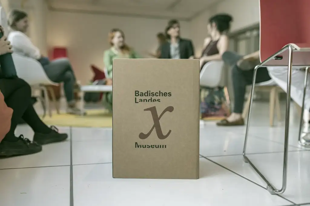 Menschen sitzen im Halbkreis und unterhalten sich, im Vordergrund eine Kiste mit der Aufschrift museum x. © Badisches Landesmuseum - Foto ARTIS - Uli Deck
