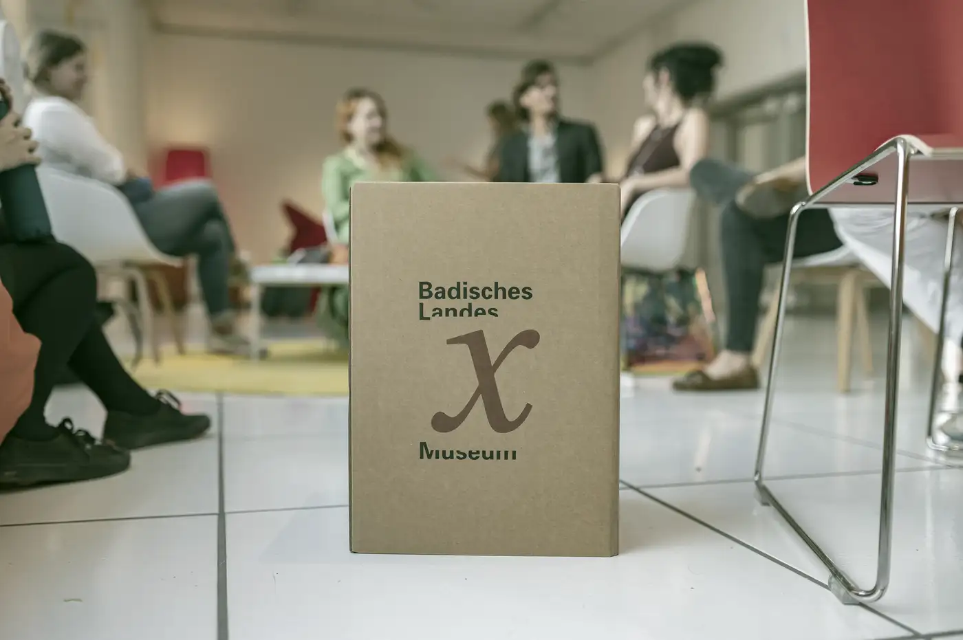 Menschen sitzen im Halbkreis und unterhalten sich, im Vordergrund eine Kiste mit der Aufschrift museum x. © Badisches Landesmuseum - Foto ARTIS - Uli Deck