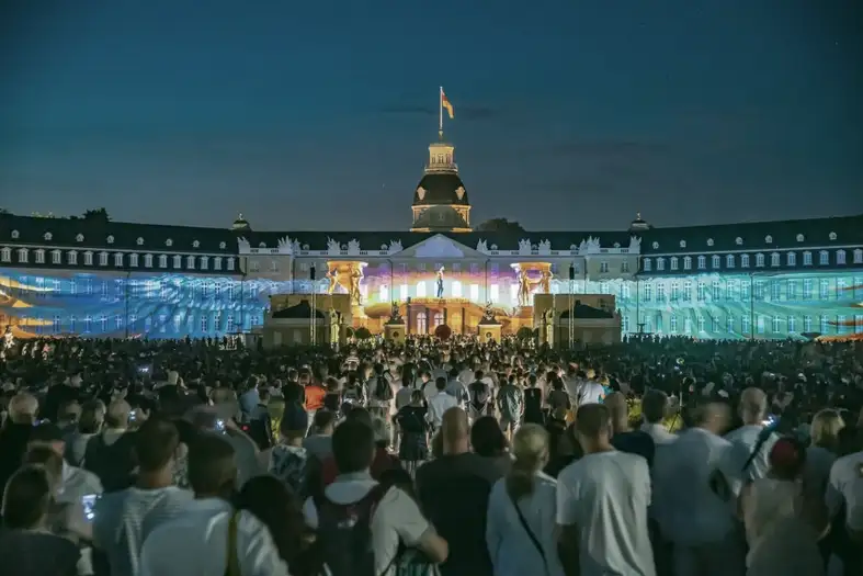 Abgebildet ist das Karlsruher Schloss. Der Himmel ist dunkelblau. Auf die Fassade des Karlsruher Schlosses sind bunte Bilder projiziert. Vor dem Schloss stehen hunderte von Menschen, die die Projektionen auf der Fassade beobachten.